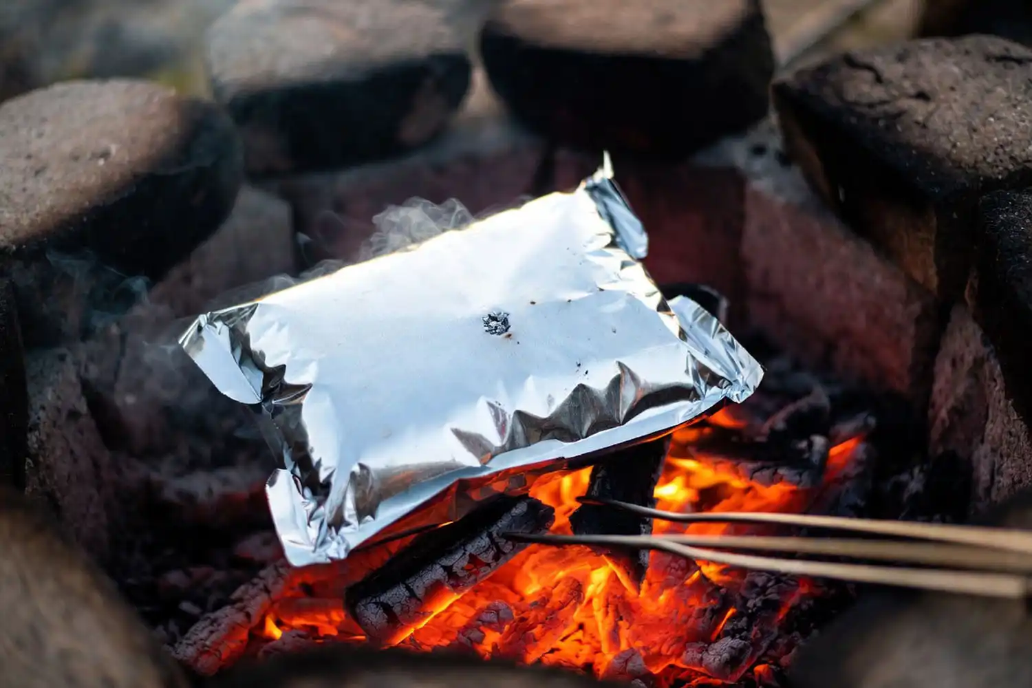 Heavy duty aluminum foil packet with chicken and potato resting on glowing campfire coals
