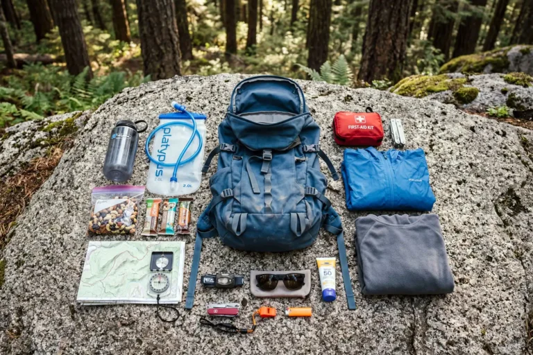 A hiking day pack with the ten essentials neatly laid out on a flat rock at a forest trailhead