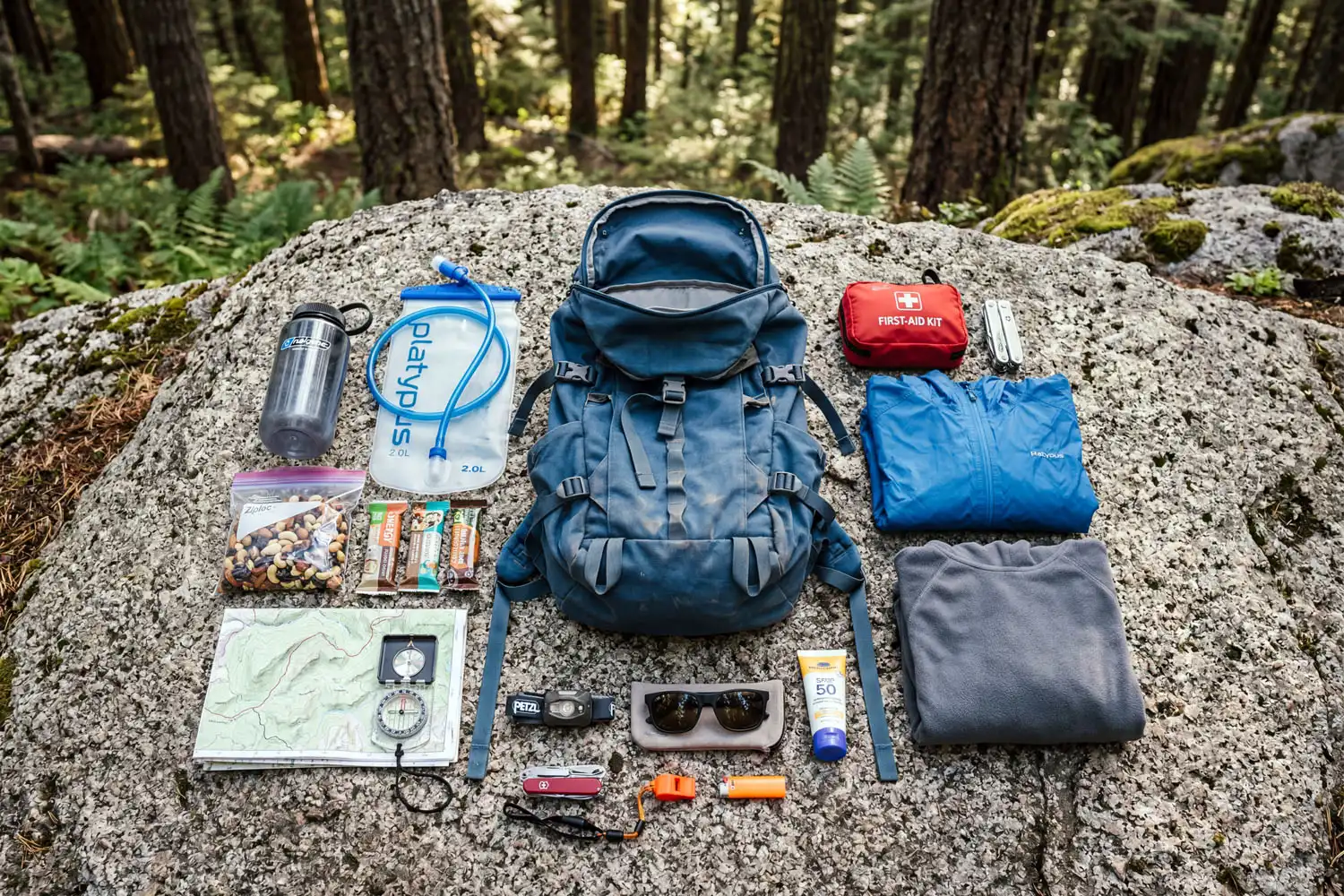 A hiking day pack with the ten essentials neatly laid out on a flat rock at a forest trailhead