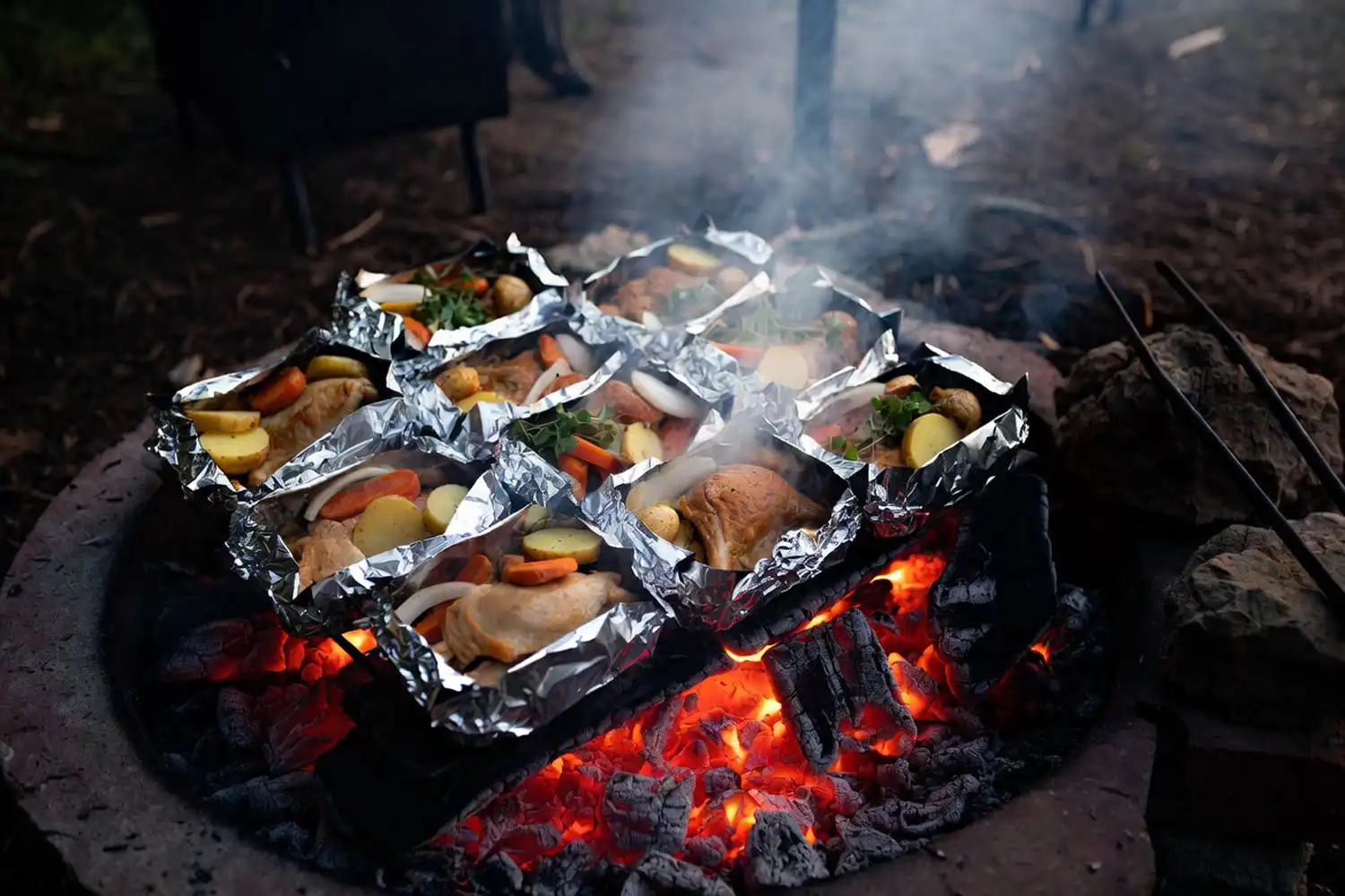Aluminum foil packet meals arranged on glowing orange and red campfire coals with smoke and steam rising in the evening